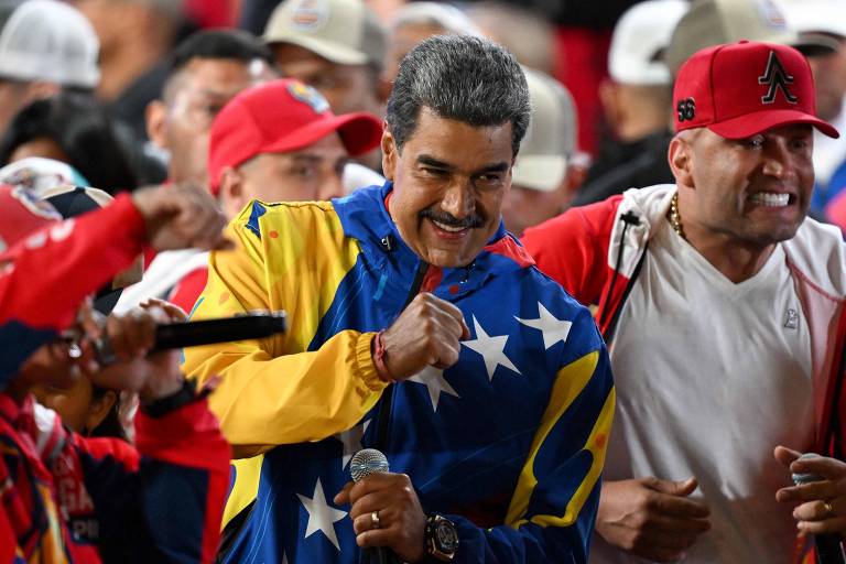 Homem de bigode e cabelo escuros agita os punhos cerrados no ar, entre apoiadores num palanque. Ele usa um abrigo esportivo que lembra a bandeira da Venezuela, azul, amarelo, vermelho e com estrelas brancas.