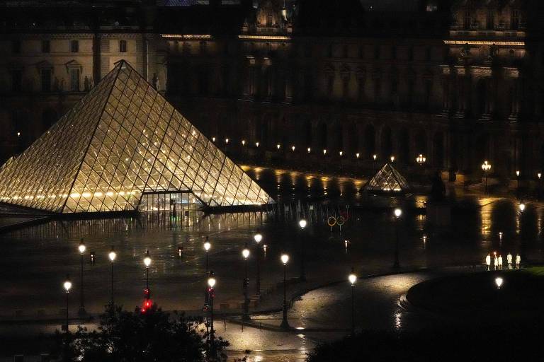 A imagem mostra a pirâmide de vidro do Museu do Louvre iluminada à noite, refletindo luzes no chão molhado. Ao fundo, o edifício do Louvre é visível, com algumas luzes acesas. Há também uma pequena pirâmide ao lado e algumas pessoas caminhando na área, além de postes de luz ao redor.
