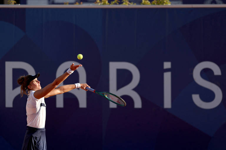 Uma jogadora de tênis está realizando um saque, com a bola sendo lançada para o alto. Ela usa uma camiseta branca e uma saia escura, e está posicionada em uma quadra de tênis. Ao fundo, há uma parede com a palavra 'PARIS' em letras grandes e estilizadas.
