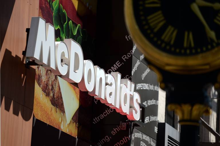 A imagem mostra o letreiro do McDonald's em destaque, com letras brancas em um fundo que exibe uma imagem de um hambúrguer com queijo. Ao fundo, há um painel preto com várias palavras relacionadas a experiências e sensações. Um relógio decorativo em primeiro plano está desfocado à direita.
