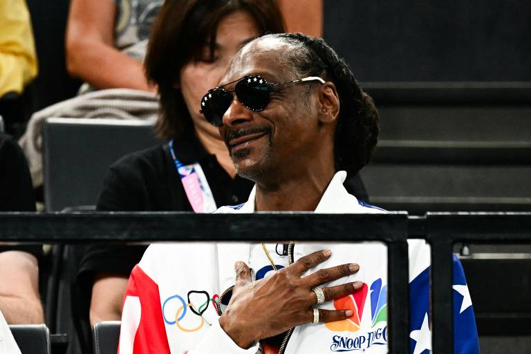 Um homem sentado em uma arquibancada, usando óculos escuros e uma jaqueta com cores e símbolos olímpicos. Ele está sorrindo e com a mão no peito, enquanto observa o evento. Ao fundo, uma mulher está parcialmente visível.
