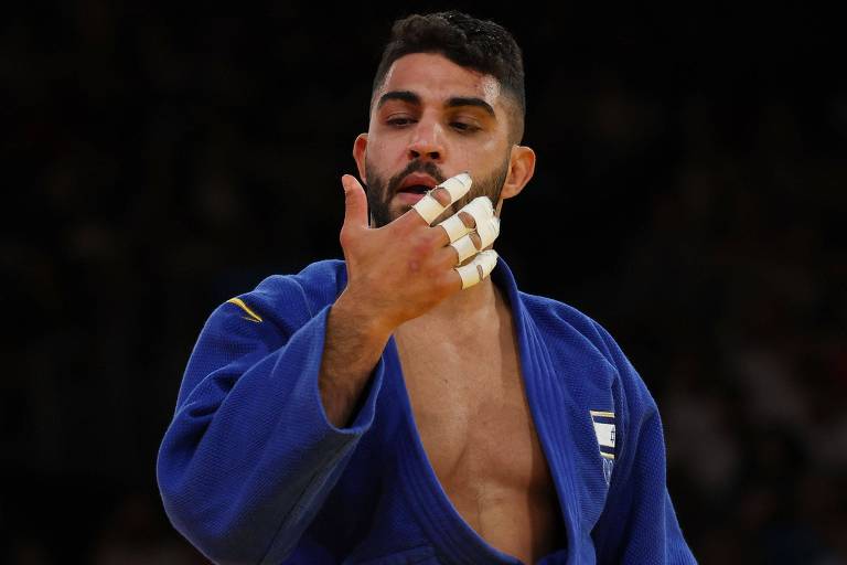 Um atleta de judô está em uma competição, vestindo um quimono azul. Ele está olhando para a mão direita, que tem os dedos enfaixados. O fundo é desfocado, sugerindo um ambiente de competição.
