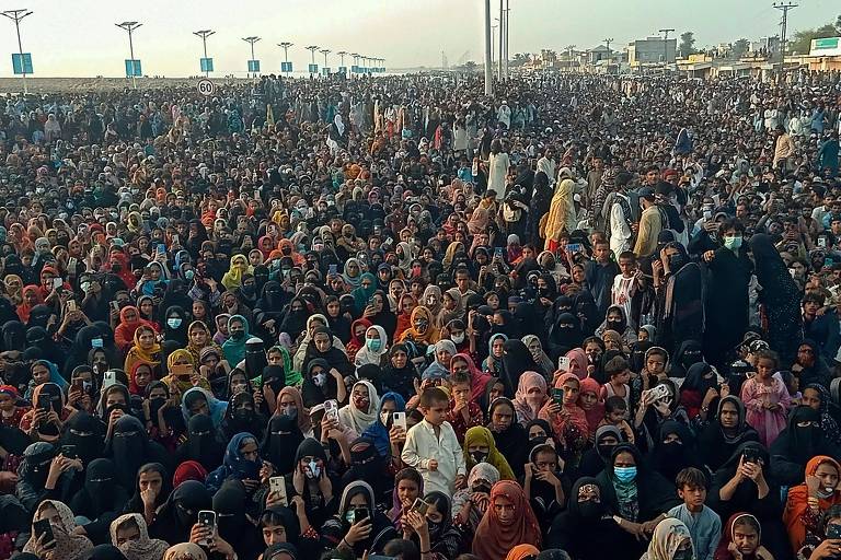 A imagem mostra uma grande multidão reunida em um evento ao ar livre. As pessoas estão vestidas com roupas de diversas cores e estilos， muitas usando lenços ou coberturas na cabeça. O ambiente parece ser um espaço amplo， com estruturas ao fundo e uma iluminação natural. A multidão é densa e parece estar atenta a algo que está acontecendo à frente