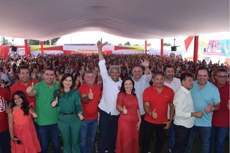 A imagem mostra um grupo de pessoas posando para a foto em um evento político. No primeiro plano, há várias pessoas vestindo camisas de diferentes cores, principalmente vermelho e verde, algumas levantando o polegar. Ao fundo, uma grande multidão está reunida sob uma tenda, com bandeiras e cartazes visíveis. O ambiente parece festivo e organizado.