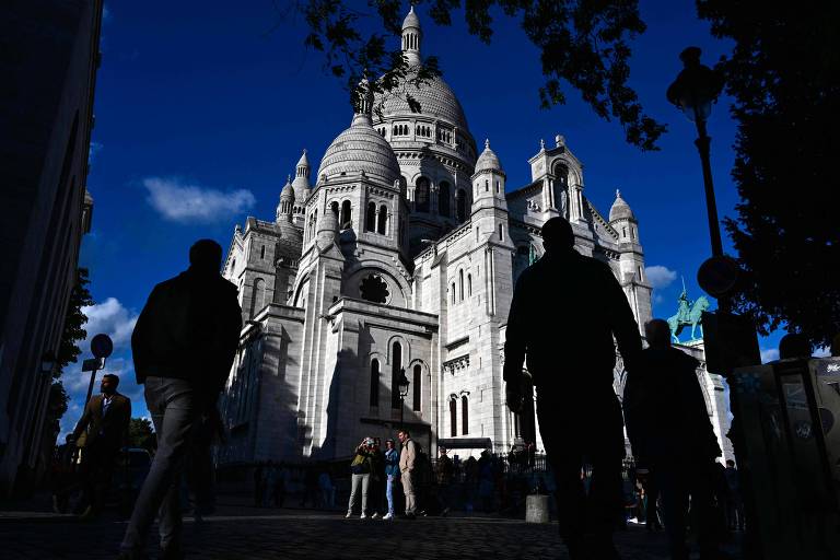 A imagem mostra a Basílica de Sacré-Cœur em Paris, com um céu azul ao fundo e nuvens brancas. Silhuetas de pessoas caminham em primeiro plano, algumas paradas, enquanto outras tiram fotos. A estrutura da basílica é visível, destacando suas cúpulas e detalhes arquitetônicos. Há também um poste de luz e uma estátua visível à direita.
