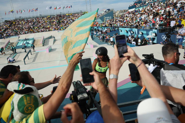A imagem mostra uma multidão em um evento de skate, com várias pessoas levantando celulares e uma bandeira. No fundo, há uma pista de skate e uma grande quantidade de espectadores assistindo ao evento. Ao centro, uma atleta com uniforme amarelo do Brasil e capacete. A atmosfera é de entusiasmo e apoio.