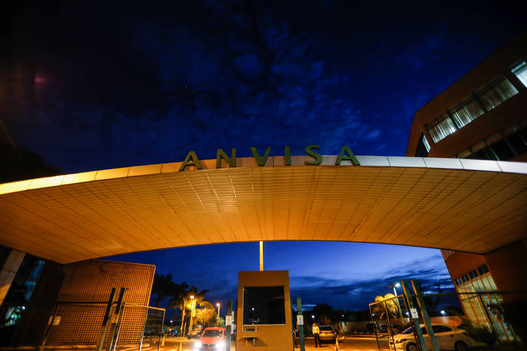 A imagem mostra a entrada da ANVISA (Agência Nacional de Vigilância Sanitária) iluminada à noite. O céu está escuro com nuvens, e a estrutura da entrada é moderna, com um arco que exibe o nome 'ANVISA' em letras grandes e iluminadas. Há uma portaria visível e iluminação ao redor.