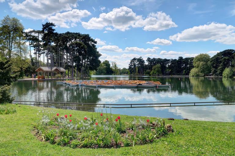 A imagem mostra um lago tranquilo cercado por árvores altas e um céu azul com nuvens brancas. Na parte inferior, há um canteiro de flores coloridas em um gramado verde. Ao fundo, uma pequena ilha no lago é visível, refletindo a paisagem ao redor.