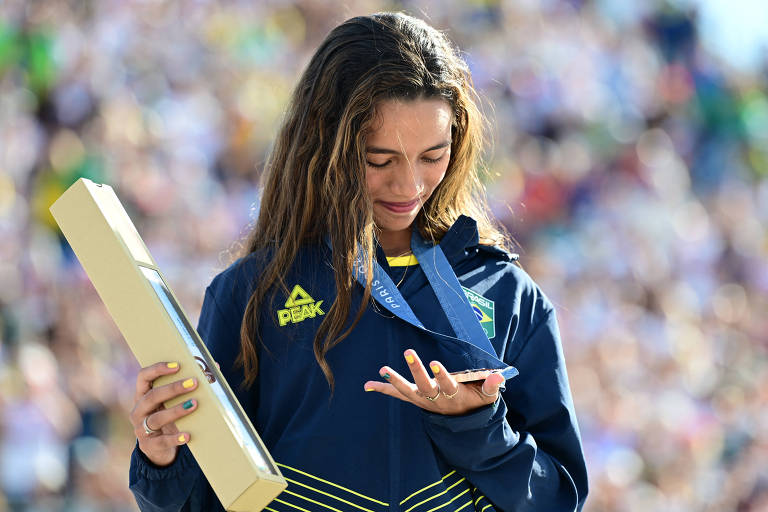 Uma atleta está em um pódio, segurando uma caixa e olhando para uma medalha em sua mão. Ela parece emocionada, com lágrimas nos olhos, vestindo uma jaqueta azul com detalhes em amarelo. Ao fundo, há uma multidão assistindo ao evento.
