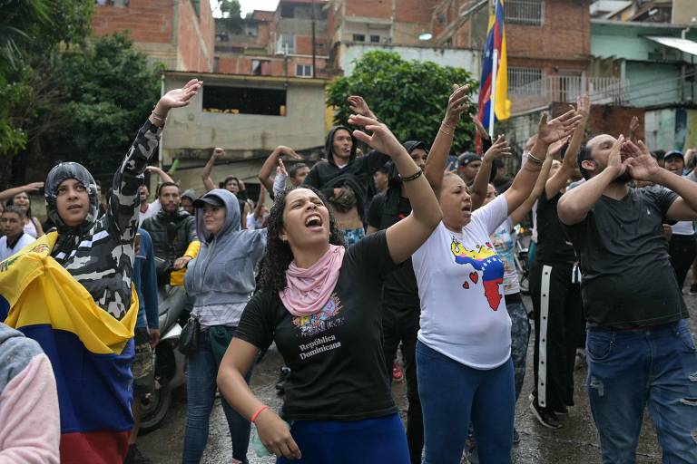 A imagem mostra um grupo de pessoas participando de uma manifestação nas ruas. Algumas pessoas estão levantando os braços e expressando emoções, enquanto outras seguram bandeiras da Venezuela. O ambiente é urbano, com prédios ao fundo e um clima de agitação e apoio. A maioria das pessoas está vestida de maneira casual, e algumas usam roupas com as cores da bandeira venezuelana.