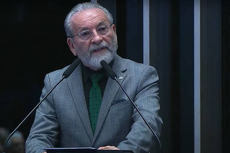 Um homem de cabelo grisalho e barba, vestido com um terno cinza e uma gravata verde, está falando em um microfone. Ele parece estar em um ambiente formal, possivelmente em um evento ou sessão legislativa, com um fundo de painéis verticais em tons escuros.