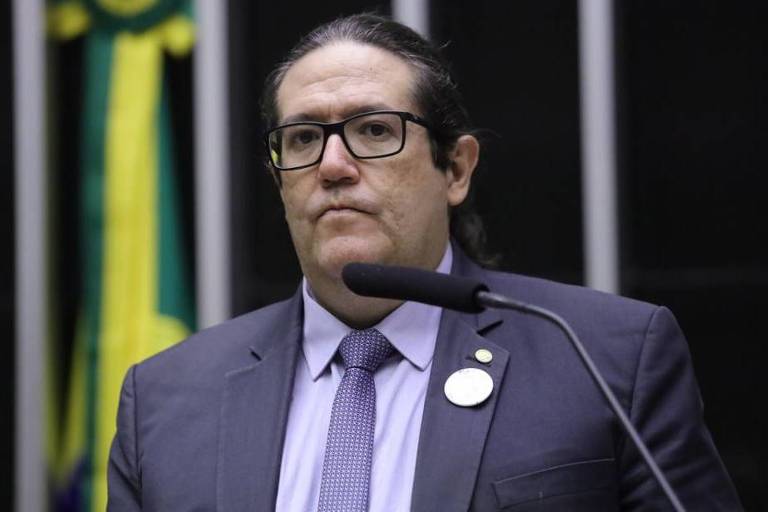 Homem branco, de óculos com armações pretas, terno azul marinho, gravata da mesma cor, em tom um pouco mais claro, discursa em tribuna.