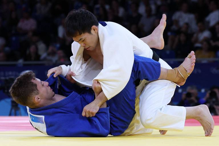 Vestindo quimono branco, o japonês Ryuju Nagayama se impõe ao brasileiro Michel Augusto, que veste azul e está deitado de costas no tatame, em combate do judô na categoria até 60 quilos nas Olimpíadas de Paris