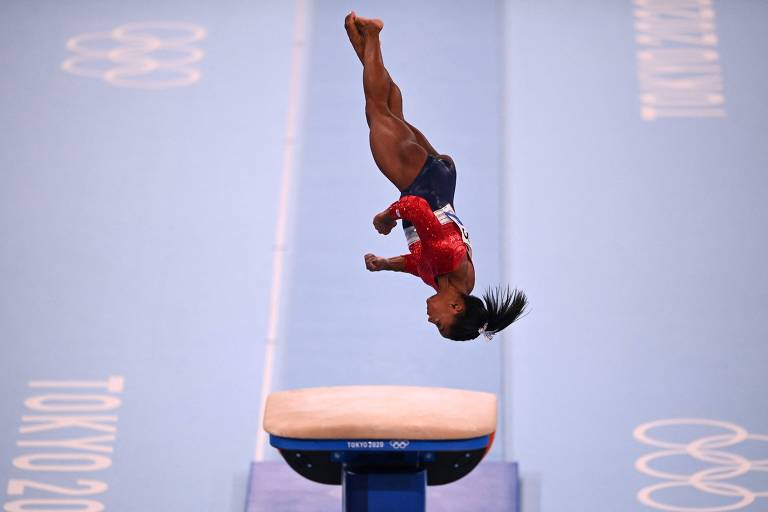 Uma ginasta está realizando um salto sobre um aparelho de salto, com o corpo em posição invertida. Ela usa um uniforme vermelho e azul, e seu cabelo está preso em um coque. O fundo é uma superfície azul com o logotipo dos Jogos Olímpicos de Tóquio 2020.
