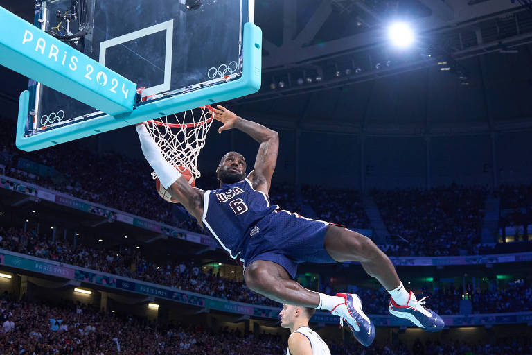 Um jogador de basquete está realizando uma enterrada em uma quadra iluminada. Ele está vestindo um uniforme azul e branco, com o número 8 visível. O aro da cesta está em destaque, e o jogador está suspenso no ar, com uma expressão de concentração. Ao fundo, há uma multidão assistindo ao jogo.
