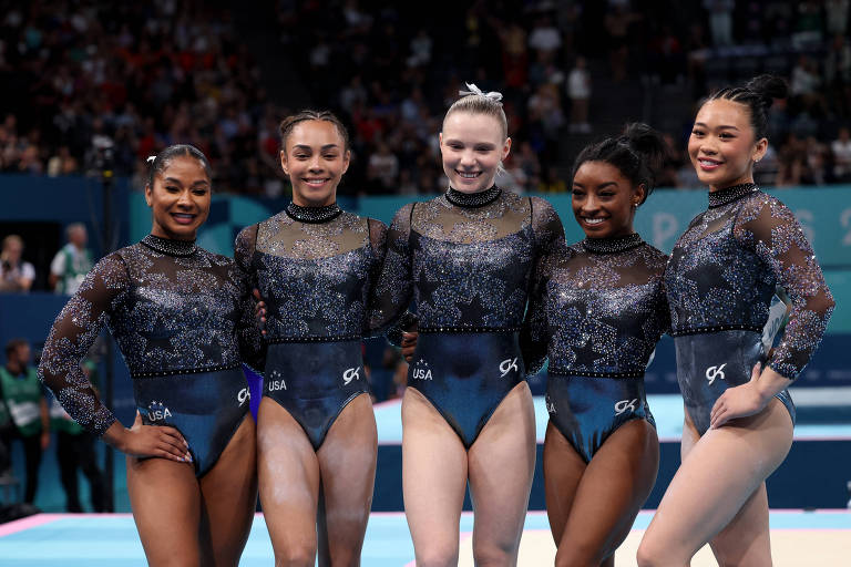 Cinco ginastas estão posando juntas em um evento de ginástica. Elas vestem uniformes de competição com detalhes brilhantes e estão sorrindo para a câmera. O fundo mostra uma multidão assistindo ao evento.
