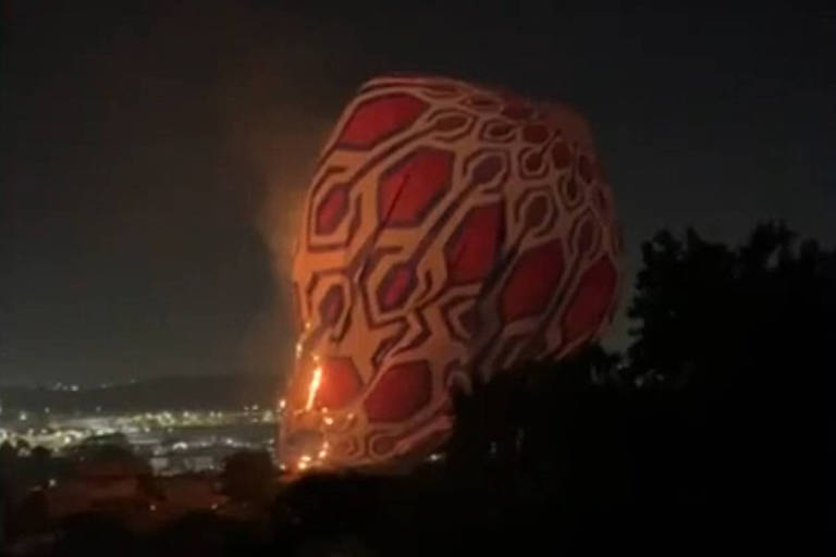 A imagem mostra um balão de ar quente iluminado à noite, com um padrão vermelho e branco. O balão está parcialmente inflado e em chamas, emitindo uma luz laranja, e caído sobre cópa de árvores. Ao fundo, é possível ver uma cidade iluminada.