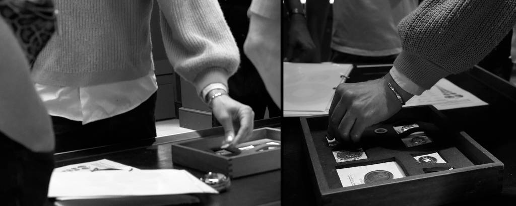 A imagem em preto e branco mostra duas mãos interagindo com uma caixa de madeira em uma mesa. À esquerda, uma mão está segurando um objeto, enquanto à direita, outra mão está colocando um item na caixa, que contém várias peças, possivelmente moedas ou medalhas. Ao fundo, há outras pessoas e papéis sobre a mesa.