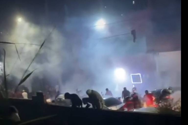 A imagem mostra uma cena noturna com fumaça densa e várias pessoas correndo em meio a escombros. Há luzes brilhantes ao fundo, possivelmente de um edifício ou veículos de emergência. A atmosfera é caótica, com pessoas se movendo 