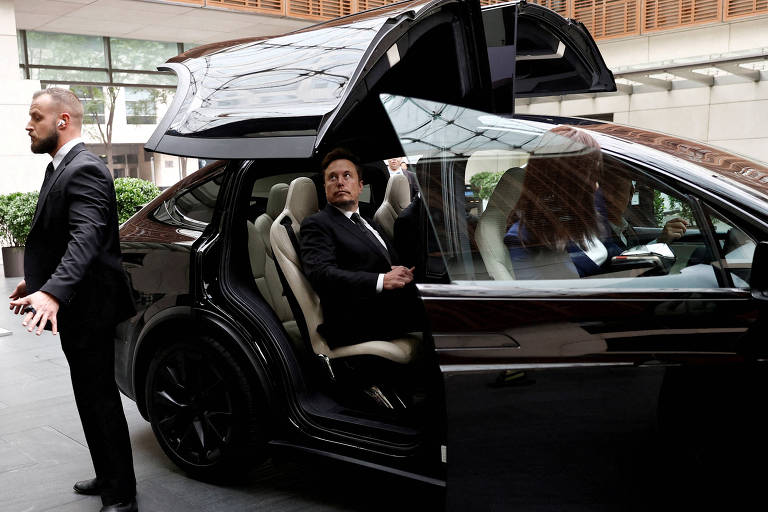 A imagem mostra um carro preto com portas que se abrem para cima, estacionado em um ambiente interno elegante. Dentro do carro, Elon Musk está sentado, enquanto dois seguranças em trajes formais estão próximos, um deles gesticulando. O ambiente ao redor é moderno, com plantas e janelas grandes ao fundo.