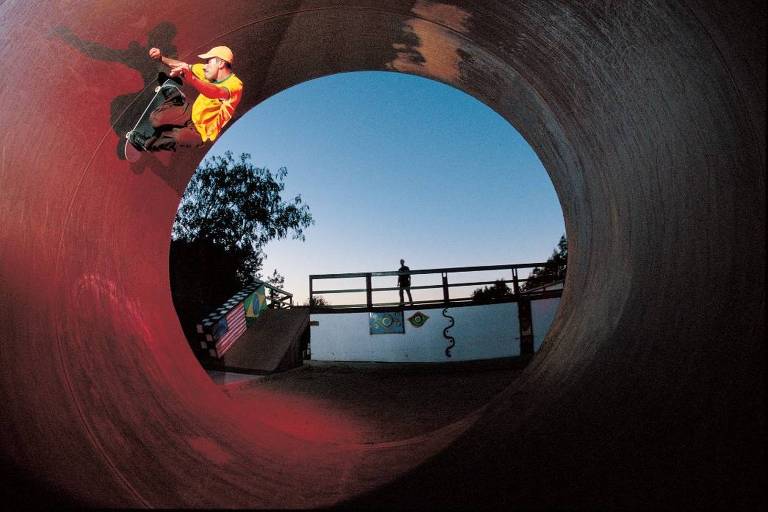 A imagem mostra um skatista realizando uma manobra dentro de um tubo de skate. O skatista está vestido com uma camiseta amarela e um boné, enquanto faz uma manobra com o skate. Ao fundo, há uma estrutura de madeira com uma pessoa em pé observando. O céu está claro, indicando o final do dia, e há árvores ao redor do local.