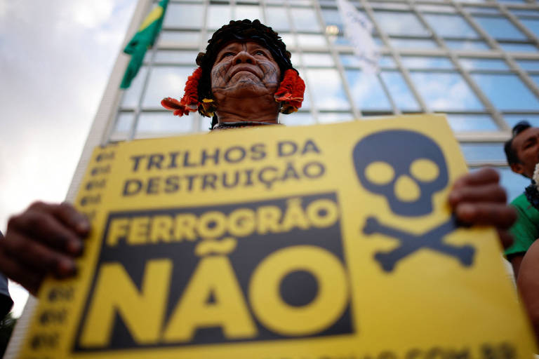 Homem segura cartaz que diz: trilhos da destruição, ferrogrão não, com um símbolo de caveira; atrás dele há um edifício e uma bandeira do Brasil em um mastro
