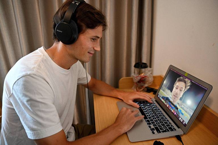 Um jovem está sentado em uma mesa, usando fones de ouvido e olhando para a tela de um laptop. Ele está sorrindo enquanto interage com o computador. Ao fundo, há cortinas e uma caneca sobre a mesa.