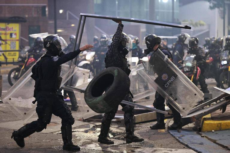 Três homens de uniforme preto e escudos transparentes retiram entulho de uma barricada