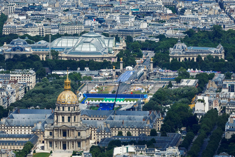 A imagem mostra uma vista aérea de Paris, destacando o domo dourado do Hôtel des Invalides em primeiro plano. Ao fundo, é possível ver várias construções, incluindo o Grand Palais e o Jardim das Tulherias, com áreas verdes visíveis entre os edifícios. O céu está claro e a cidade se estende em várias direções.