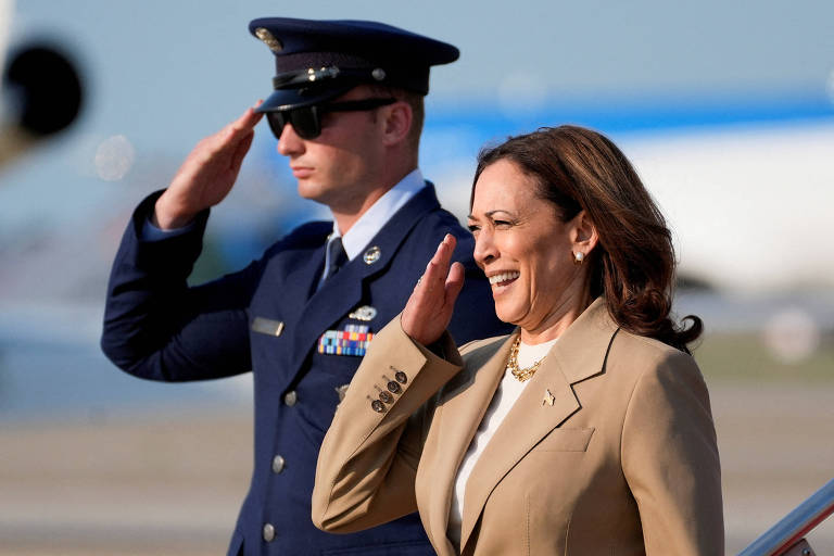 A imagem mostra uma mulher sorridente, vestindo um terno claro, acenando com a mão enquanto um homem em uniforme militar, usando óculos escuros, a saúda com a mão em posição de respeito. Ao fundo, é possível ver uma aeronave e um céu claro.