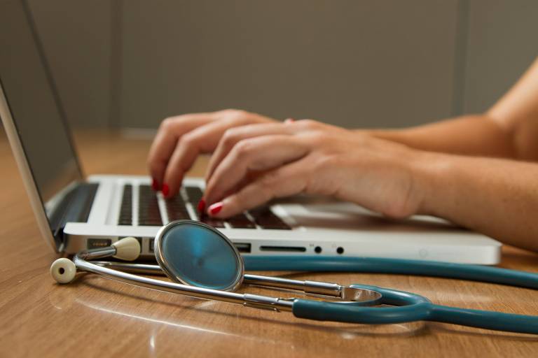 A imagem mostra um laptop em uma mesa de madeira, com as mãos de uma pessoa digitando no teclado. Ao lado do laptop, há um estetoscópio azul. O fundo é desfocado, mas sugere um ambiente de trabalho ou consultório.