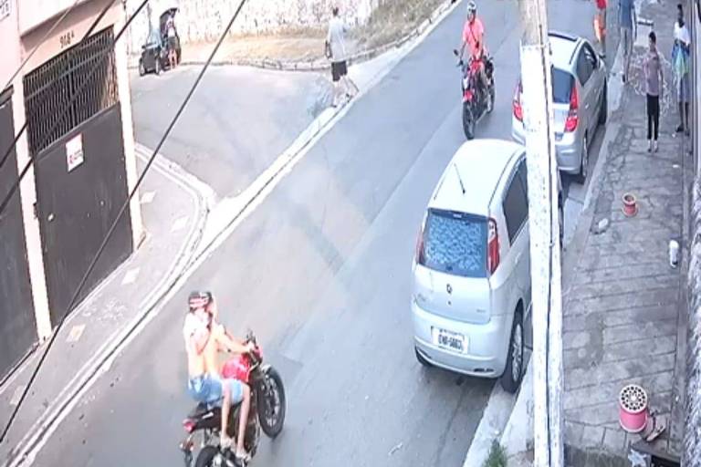 A imagem mostra uma rua com dois motociclistas. Ao fundo, há um carro prateado estacionado e algumas pessoas na calçada. Um homem está caminhando e outras pessoas estão paradas, observando a cena. A rua é pavimentada e há postes de luz e fios visíveis