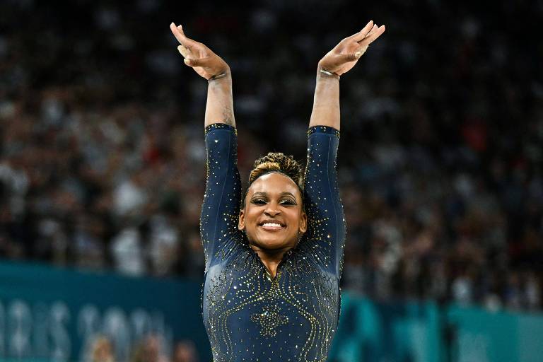 A brasileira Rebeca Andrade, vestindo collant preto, ergue os braços ao completar sua apresentação na tarde na final por equipes da ginástica artística nas Olimpíadas de Paris