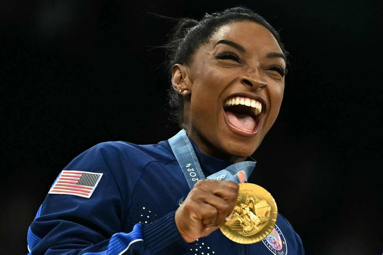Uma atleta está sorrindo amplamente enquanto segura uma medalha de ouro. Ela usa um uniforme azul com detalhes em branco e uma bandeira dos Estados Unidos no braço. O fundo é desfocado, sugerindo um ambiente de celebração.
