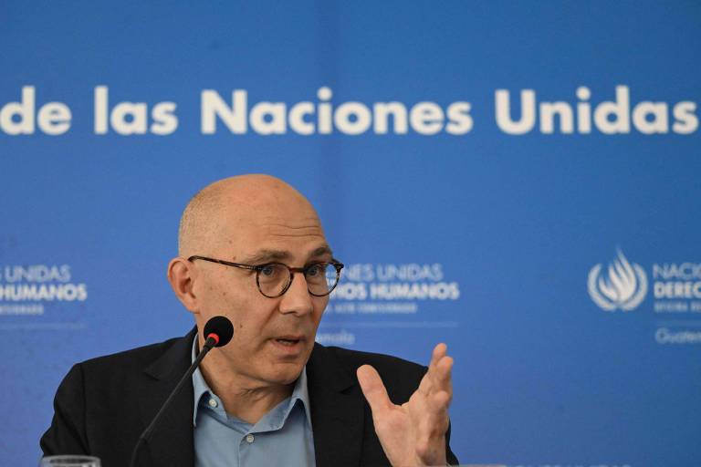 Um homem de óculos e cabelo calvo está falando em uma mesa, gesticulando com a mão. Ao fundo, há um painel azul com o texto 'Oficina de las Naciones Unidas' e o logotipo de direitos humanos.