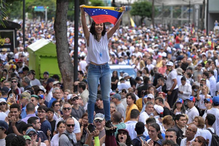 Uma multidão de pessoas participa de um protesto. No centro da imagem, uma mulher está em cima de um grupo de pessoas, segurando uma bandeira da Venezuela. A multidão é composta por pessoas de diversas idades e etnias, algumas usando bonés e camisetas brancas. Ao fundo, há árvores e uma estrutura urbana.