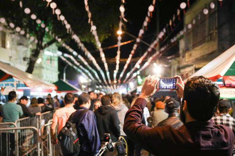 A imagem mostra uma rua movimentada à noite, decorada com luzes penduradas. Há várias barracas de comida e bebida ao longo da calçada, e um grupo de pessoas está reunido, algumas delas tirando fotos com seus celulares. A atmosfera é festiva, com muitas pessoas conversando e se divertindo.