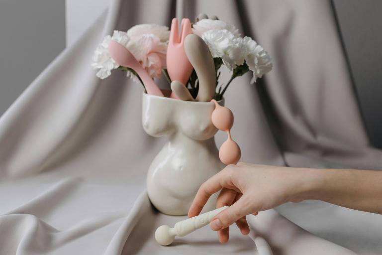 A imagem mostra um arranjo de flores brancas em um vaso que tem a forma de um torso feminino. Ao lado do vaso, há vários objetos de silicone em cores suaves, como rosa e bege, que parecem ser brinquedos de estímulo. Uma mão feminina segura um objeto branco com uma esfera na ponta, posicionado próximo ao vaso.