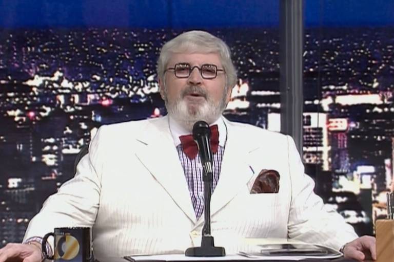 Um homem com cabelo grisalho e barba, usando um terno branco listrado e uma gravata borboleta vermelha. Ele está sentado em um cenário de programa de TV, com uma cidade iluminada ao fundo