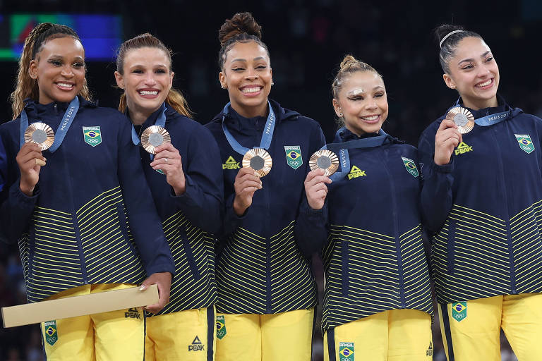 Cinco atletas brasileiras estão no pódio, sorrindo e segurando medalhas de bronze. Elas vestem uniformes amarelos e azuis, com jaquetas que têm detalhes em verde. O fundo é desfocado, sugerindo um ambiente de competição.