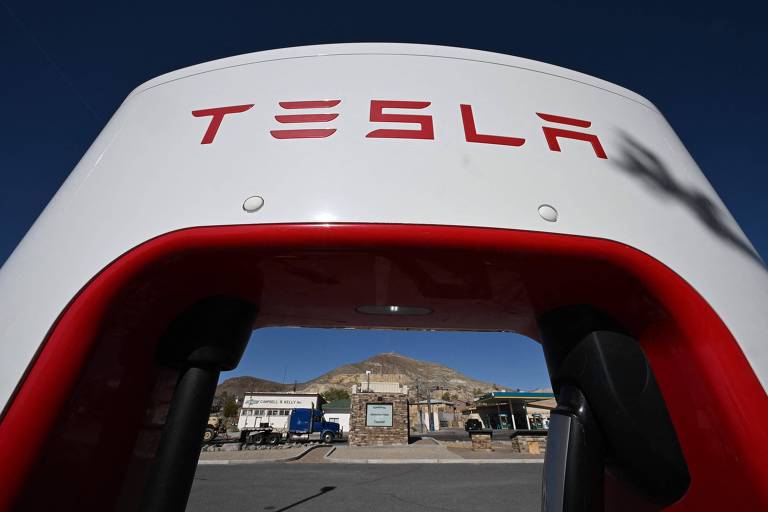 A imagem mostra uma estação de carregamento da Tesla, com a parte superior em branco e o logotipo 'TESLA' em vermelho. A vista é de dentro da estação, com um fundo que inclui uma montanha e um caminhão estacionado. O céu está claro e azul.
