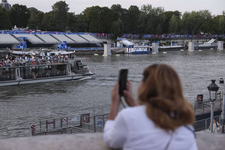 Uma mulher com cabelo castanho claro, usando uma blusa branca, está de costas, segurando um celular e tirando fotos de um barco no rio. Ao fundo, há uma multidão em um barco e estruturas ao longo da margem do rio, com árvores e um céu nublado.