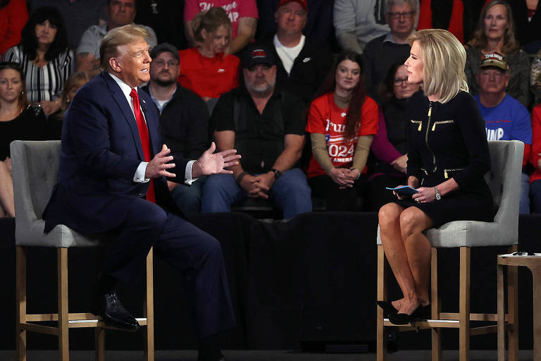 A imagem mostra Donald Trump sentado em uma cadeira, gesticulando enquanto fala. Ele está vestido com um terno escuro e uma gravata vermelha. À sua frente, uma mulher sentada em uma cadeira, segurando um bloco de notas e um microfone, parece estar fazendo perguntas. Ao fundo, uma plateia composta por várias pessoas, algumas vestindo camisetas vermelhas com a palavra 'Trump'.