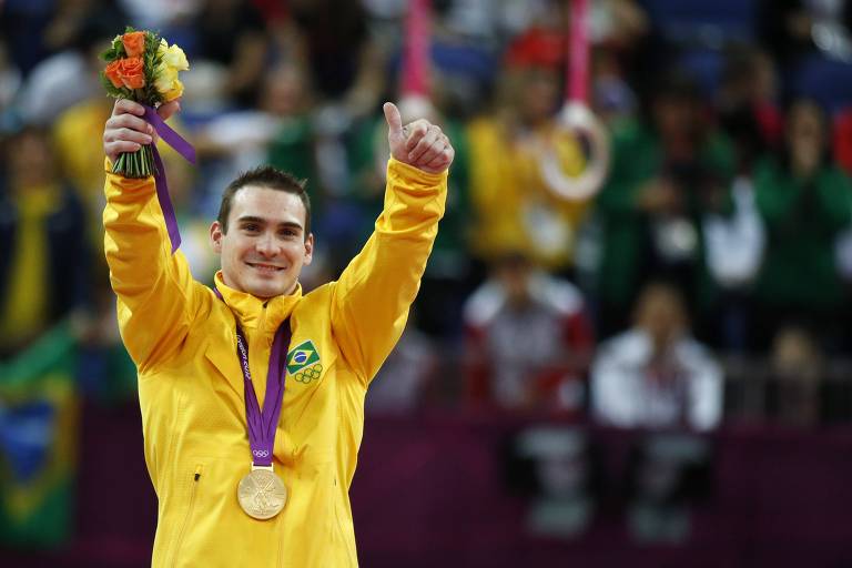 De uniforme amarelo e com flores na mão direita, o ginasta brasileiro Arthur Zanetti, com a medalha de ouro no peito, ergue os braços e sorri no pódio das Olimpíada de 2012, em Londres; ele foi campeão nas argolas