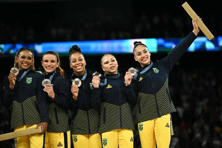 Com suas medalhas e o uniforme do Brasil, as ginastas Rebeca Andrade, Jade Barbosa, Lorrane dos Santos, Flávia Saraiva e Julia Soares celebram, sorridentes no pódio, o bronze por equipes em Paris-2024