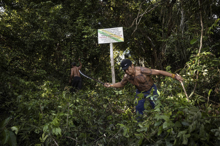 A imagem mostra dois homens trabalhando em uma área de floresta densa. Um deles está à frente, usando uma ferramenta para cortar vegetação, enquanto o outro está mais ao fundo. Ao lado deles, há uma réplica da placa de demarcação de terras indígenas. A vegetação é verde e espessa, com árvores e arbustos ao redor.