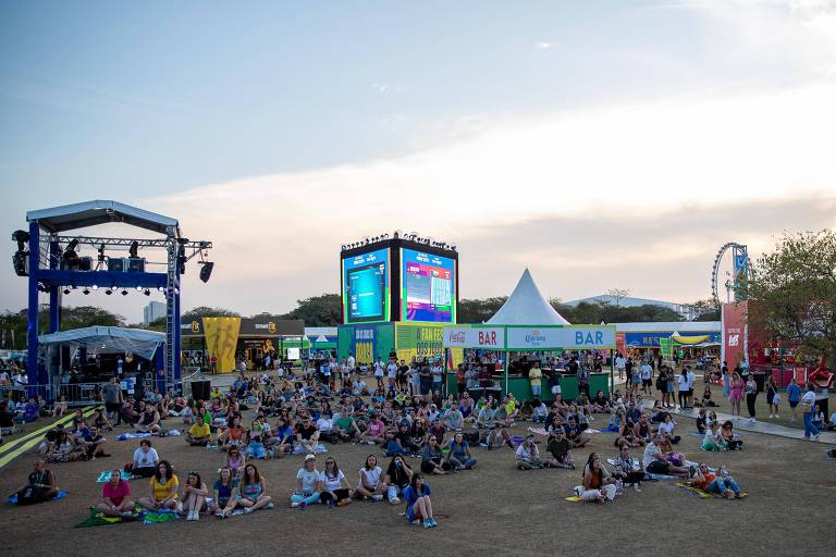 A imagem mostra um grande evento ao ar livre, com um público sentado em um gramado. No fundo, há um palco elevado com pessoas e uma tela grande exibindo informações. 