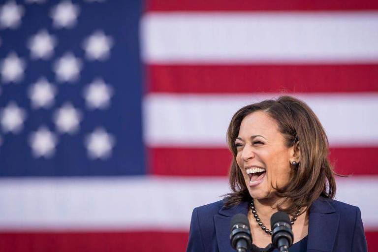 A imagem mostra uma mulher sorrindo amplamente enquanto fala em um microfone. Ao fundo, há uma grande bandeira dos Estados Unidos, com listras vermelhas e brancas e estrelas brancas em um campo azul. A mulher está vestindo um blazer escuro e tem cabelo solto.