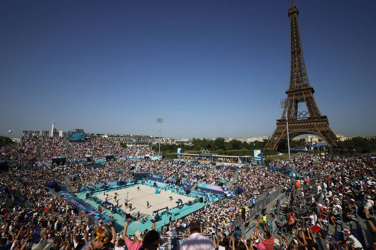 A imagem mostra uma grande multidão assistindo a uma competição de vôlei de praia em um local ao ar livre, com a Torre Eiffel visível ao fundo. O céu está limpo e ensolarado, e a quadra de vôlei é cercada por espectadores. A atmosfera é vibrante, com muitas pessoas reunidas para o evento.
