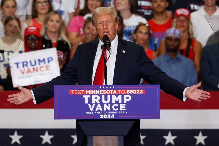 A imagem mostra um homem em um palanque, falando em um microfone. Ele está vestido com um terno escuro e uma gravata vermelha. Atrás dele, há uma multidão de apoiadores, alguns usando bonés vermelhos. Um cartaz em destaque diz 'TRUMP VANCE' e 'MAKE AMERICA GREAT AGAIN! 2024'. O fundo é decorado com uma bandeira americana.

